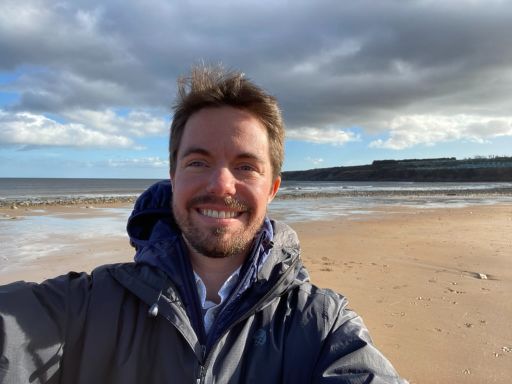 Mentor Gabe smiles for a selfie on the beach. He is wearing a winter coat, the sun is shining on his face.