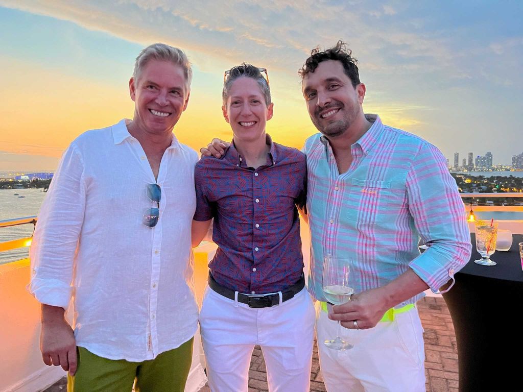 Tom, Colby, and Felipe smile at the camera while standing on a beach as the sun sets, 2022